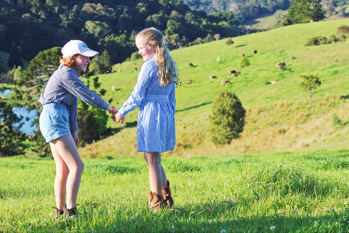 Two girls holding hands in a sunny green field with rolling hills and trees in the background.