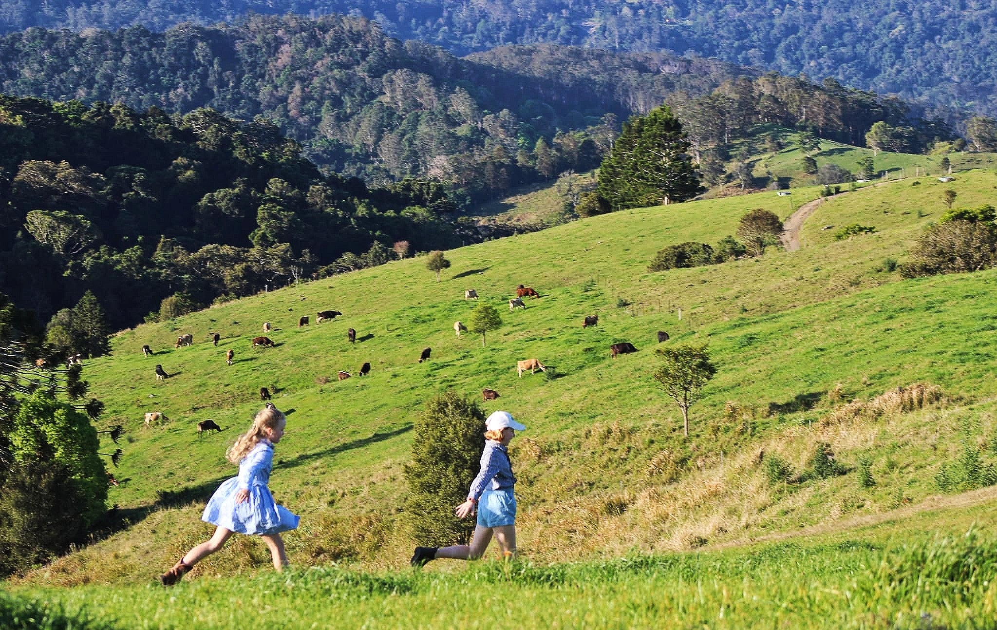 Two children in light blue dresses run across a lush green hillside with grazing cows and forested mountains in the background.
